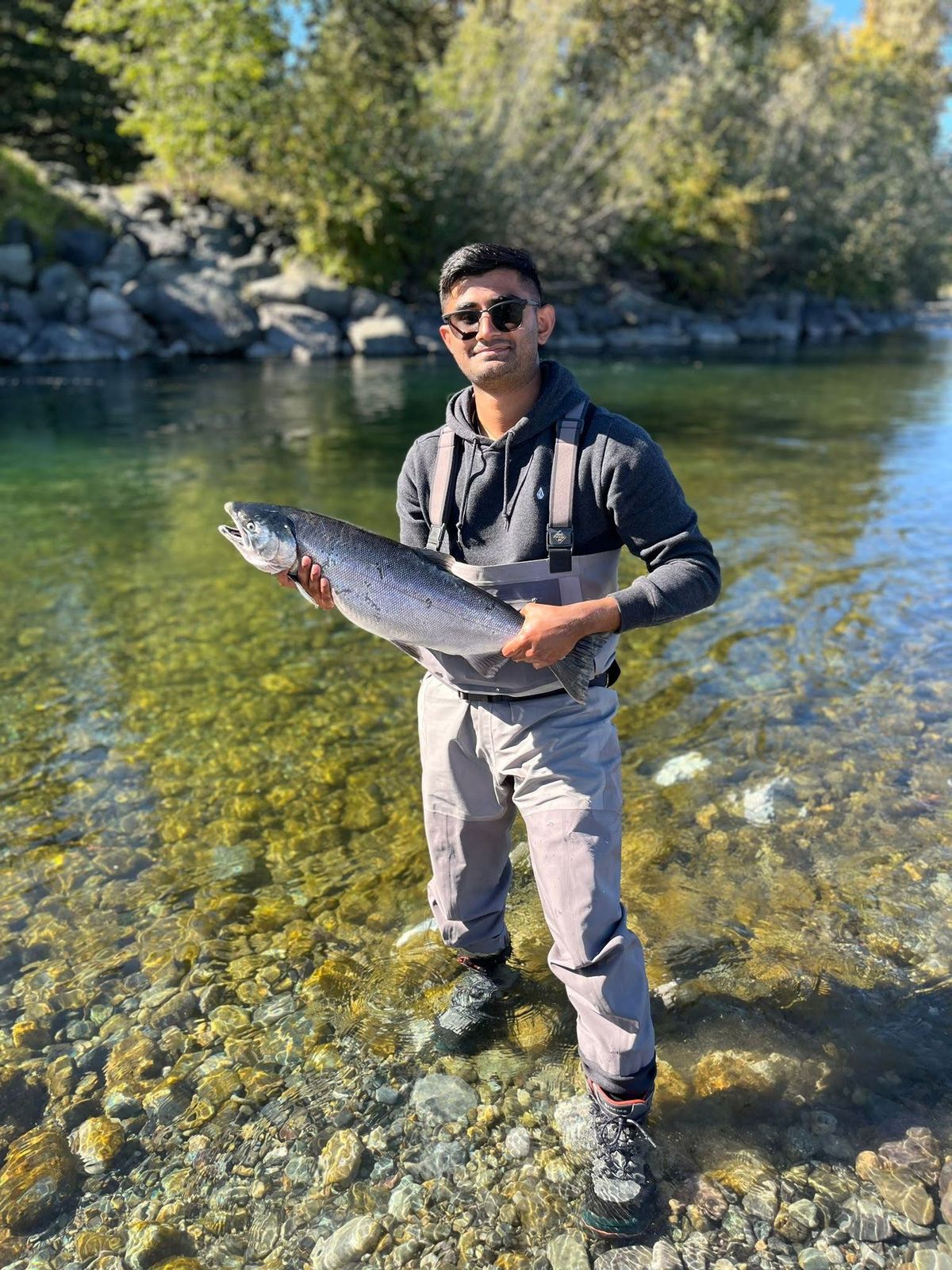 Aneesh holding a salmon in a BC river, wearing waders